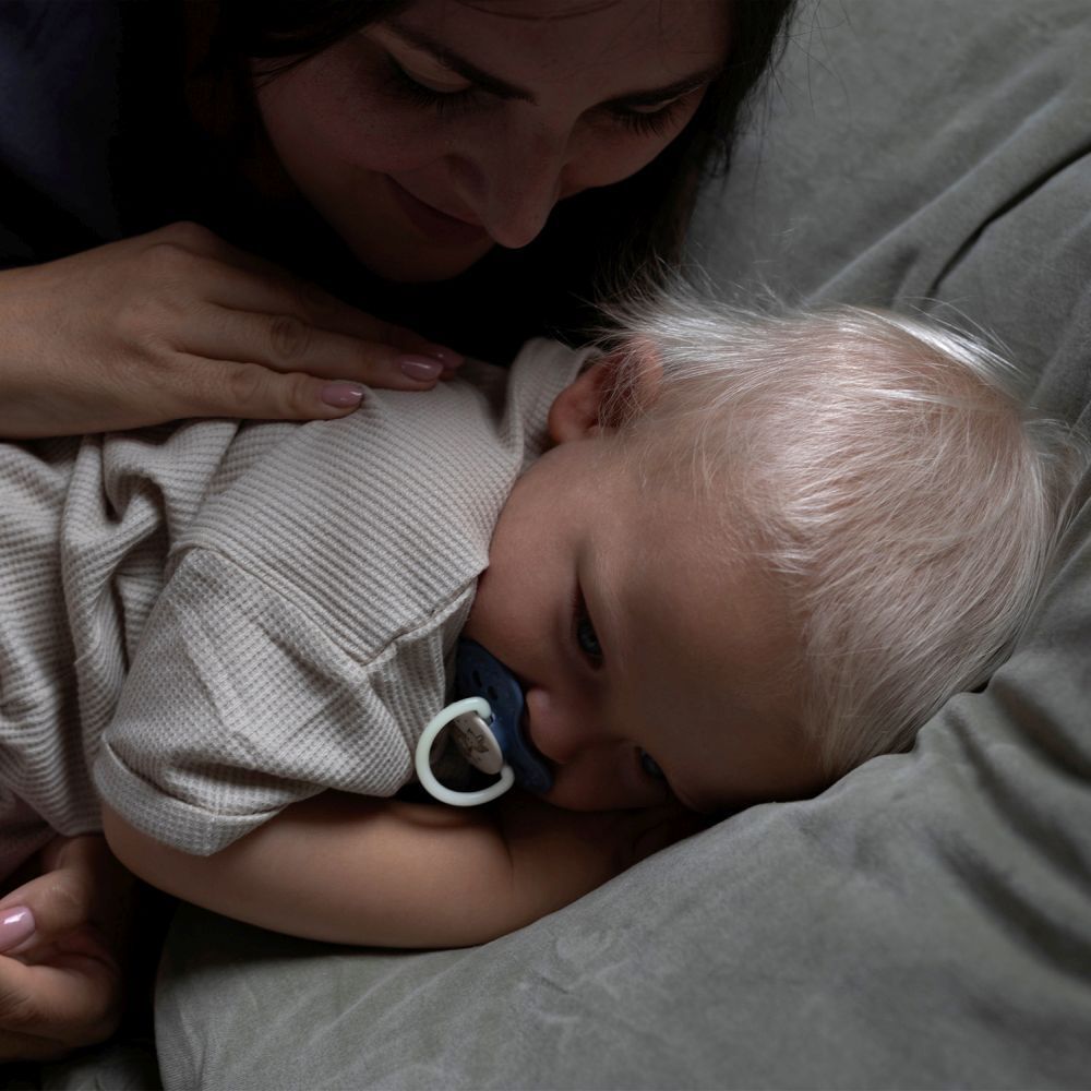 Baby ligt op een bank, fopspeen in de mond. Moeder raakt de baby aan. Fopspeen blauw.