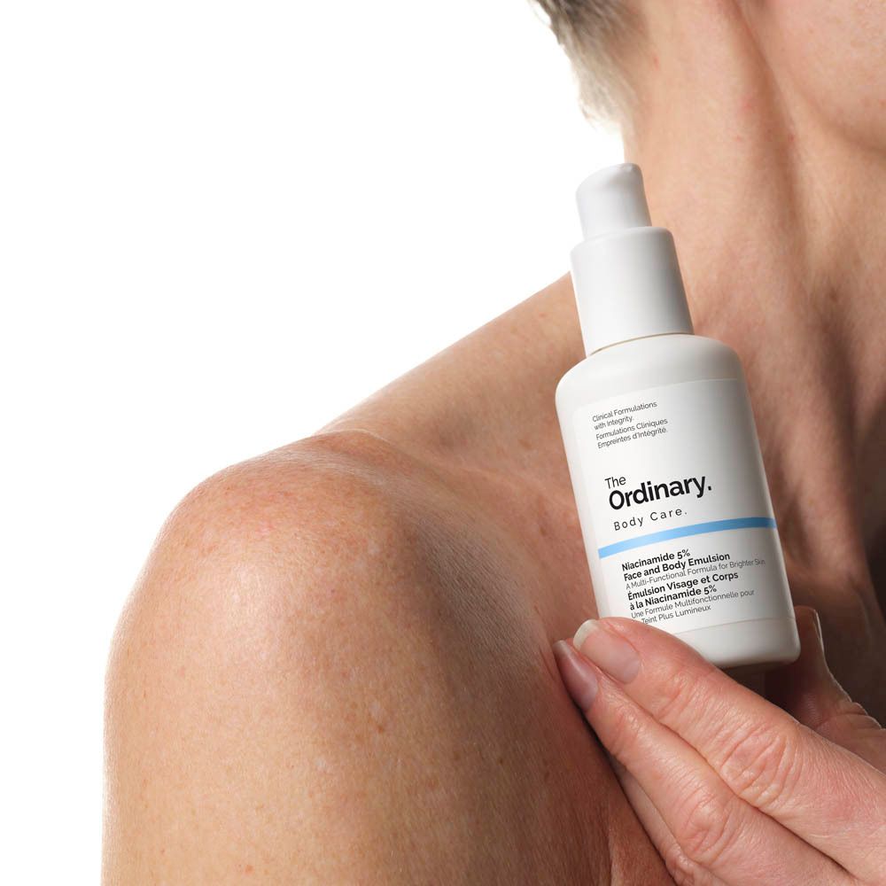 Flacon blanc avec pompe. Inscription: The Ordinary. Niacinamide 5% Face & Body Emulsion. Tenue par une personne.