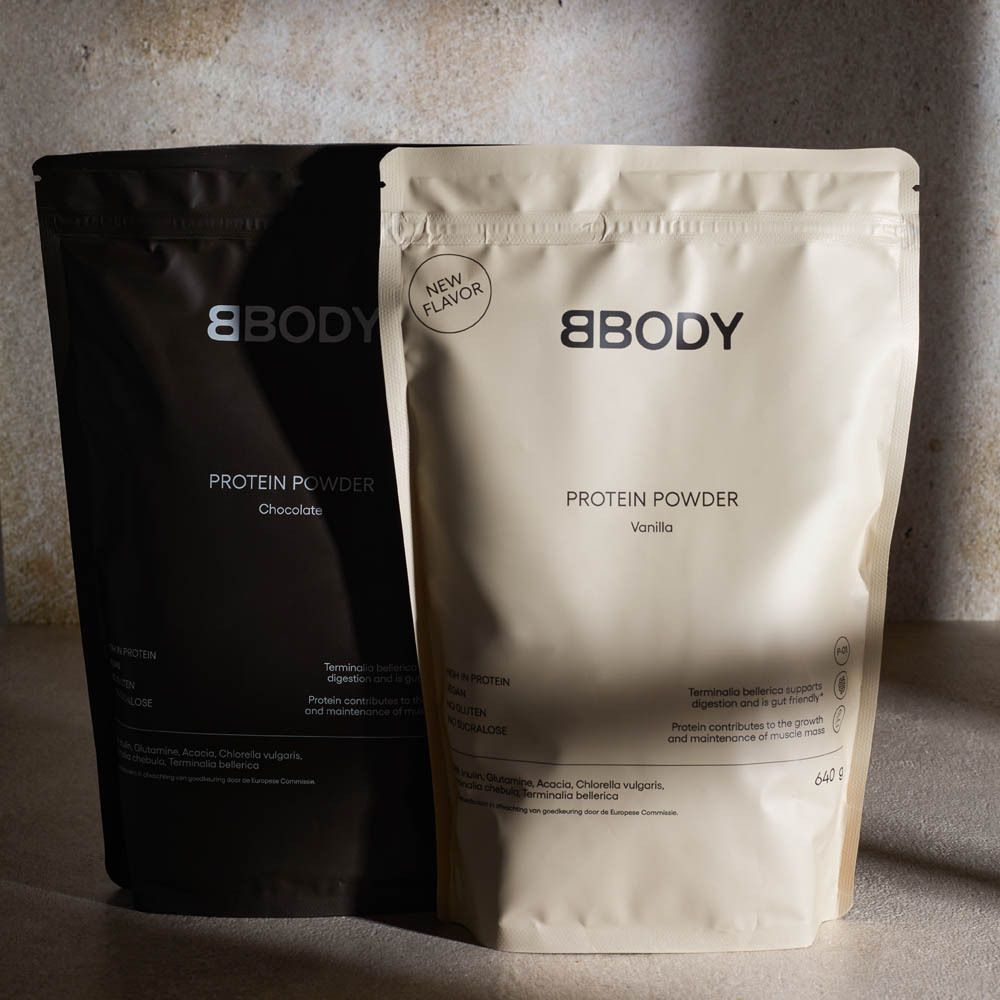 Twee BBODY Protein Powder zakken, Chocolade en Vanille. Tekst: New Flavor, 640g. Op tafel. Zwarte en beige zak.