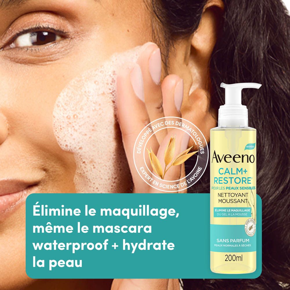 Femme se lave le visage avec Aveeno Calm+Restore Nettoyant Moussant. Flacon en arrière-plan.