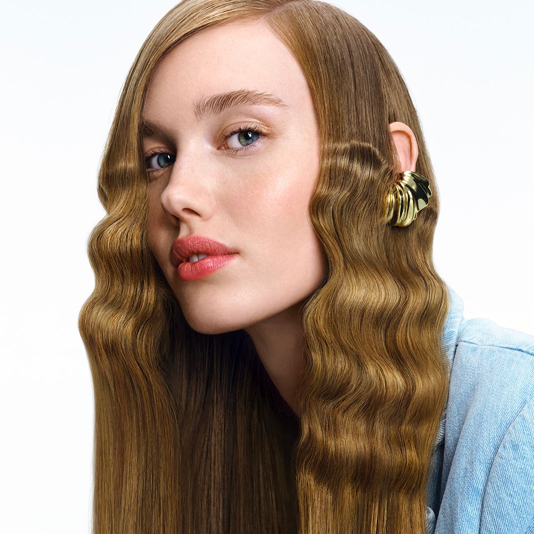 Portret van een vrouw met golvend haar. Ze draagt een gouden oorbel. Lichtblauwe jeanslook.