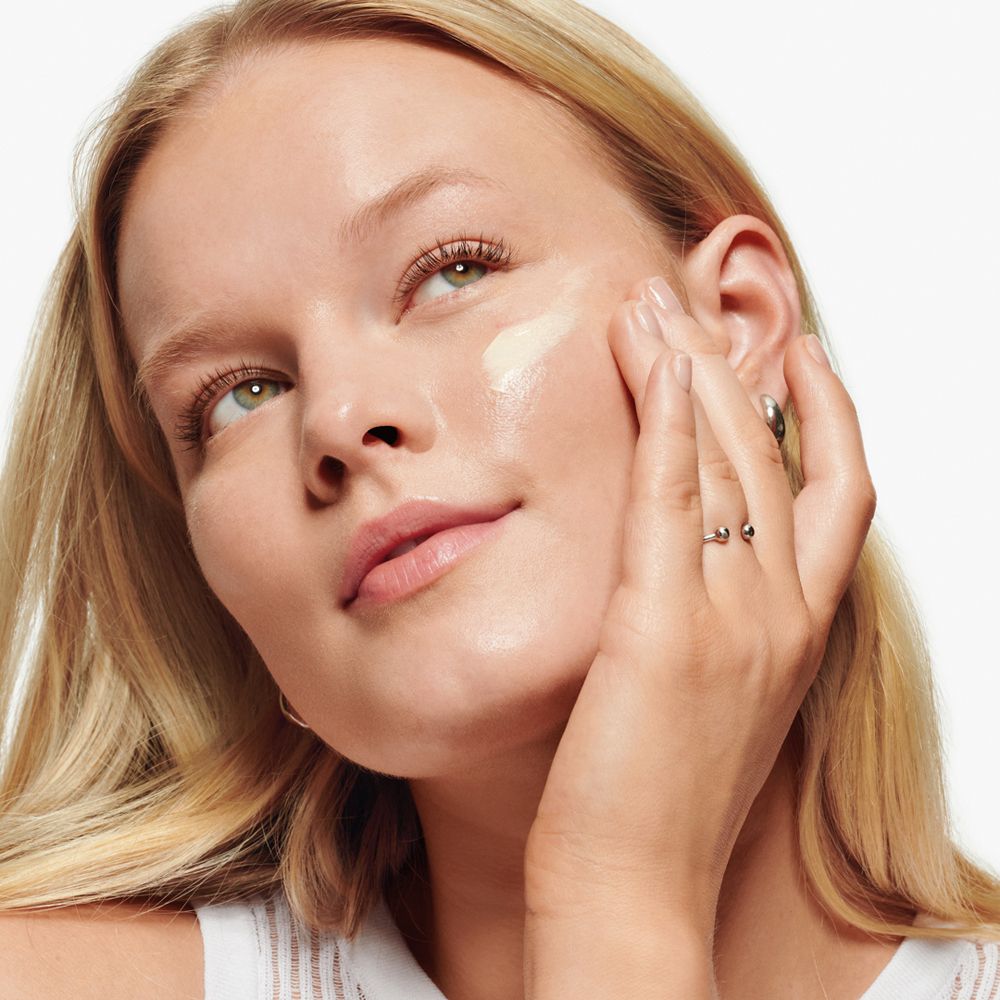Vrouw brengt crème aan op haar gezicht. Ringen aan vingers. Lichte huid, blond haar.