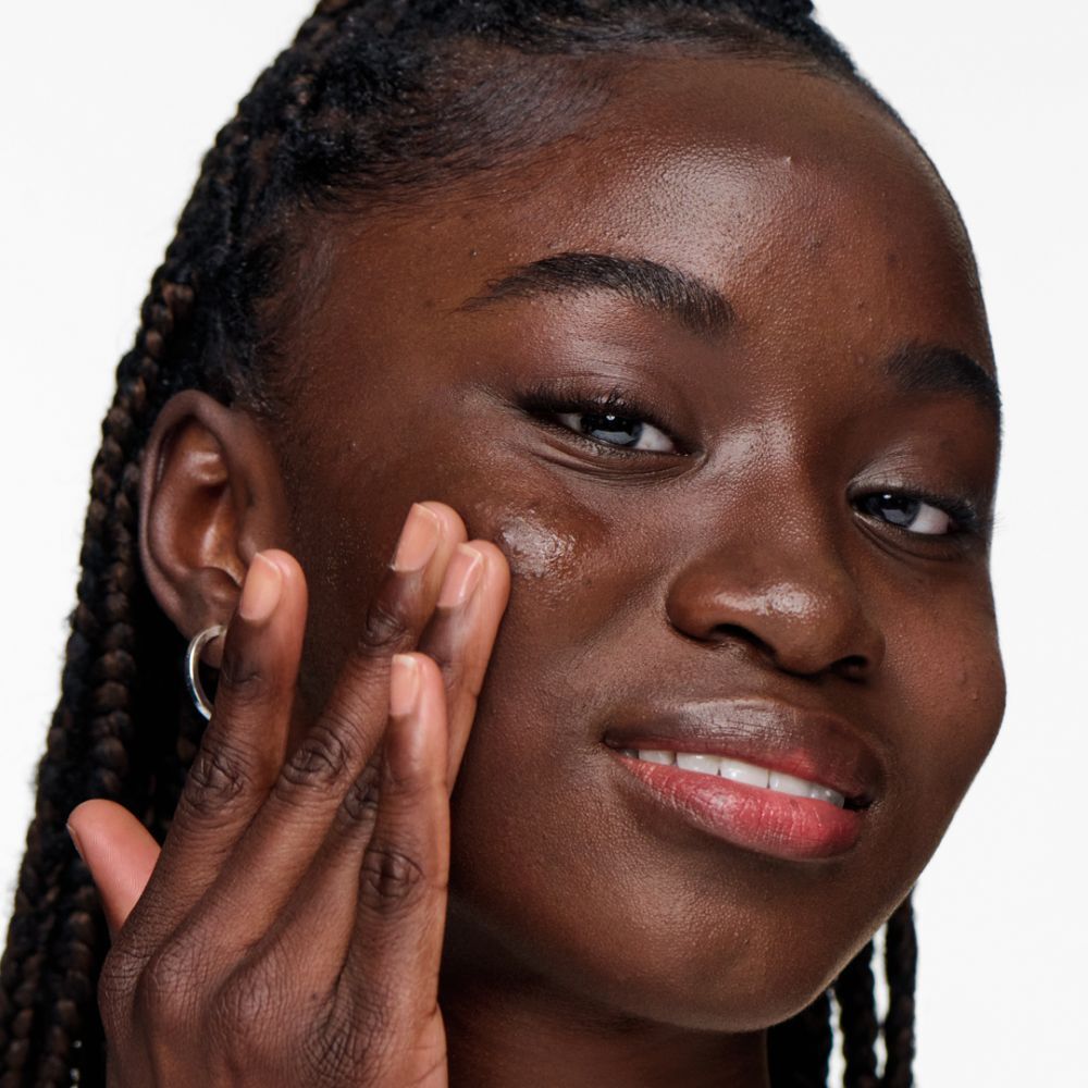 Femme à la peau foncée appliquant du gel. Elle sourit. Gel visible sur la peau.