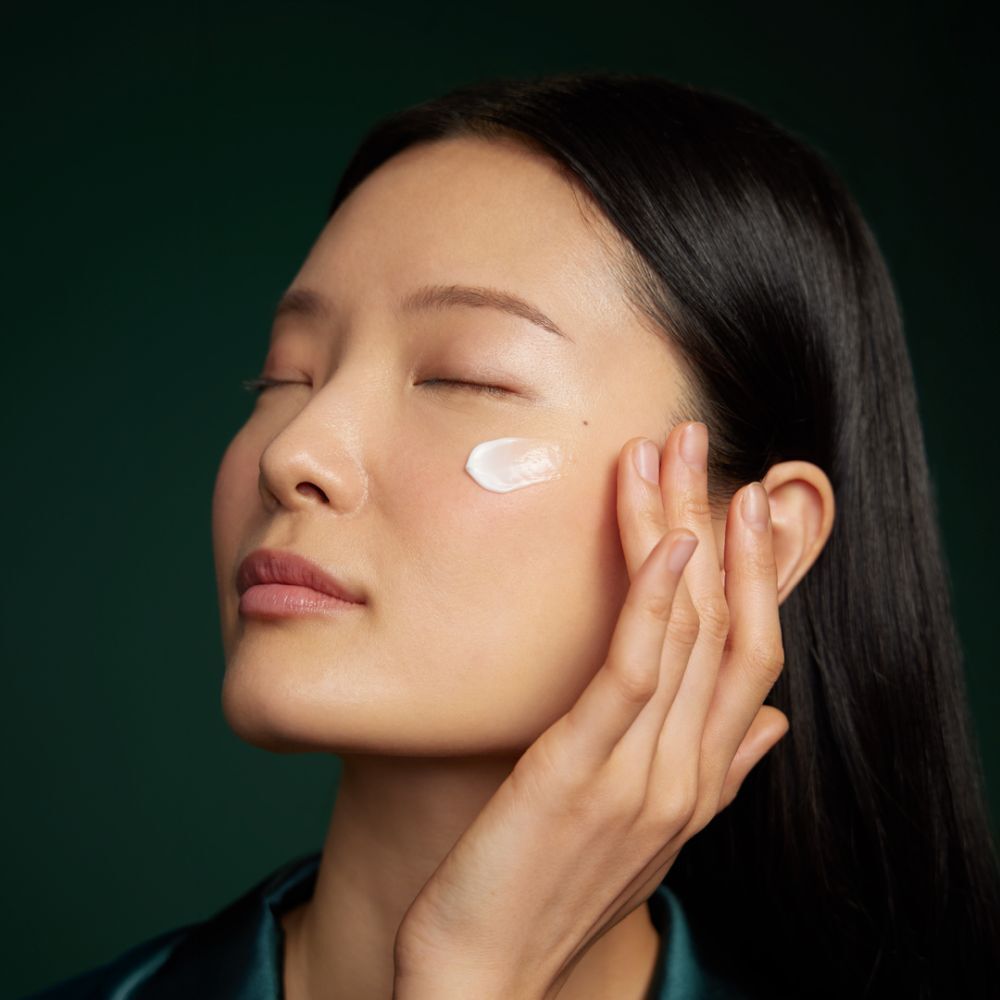 Vrouw brengt crème aan op wang. Ogen dicht, groene zijden blouse.