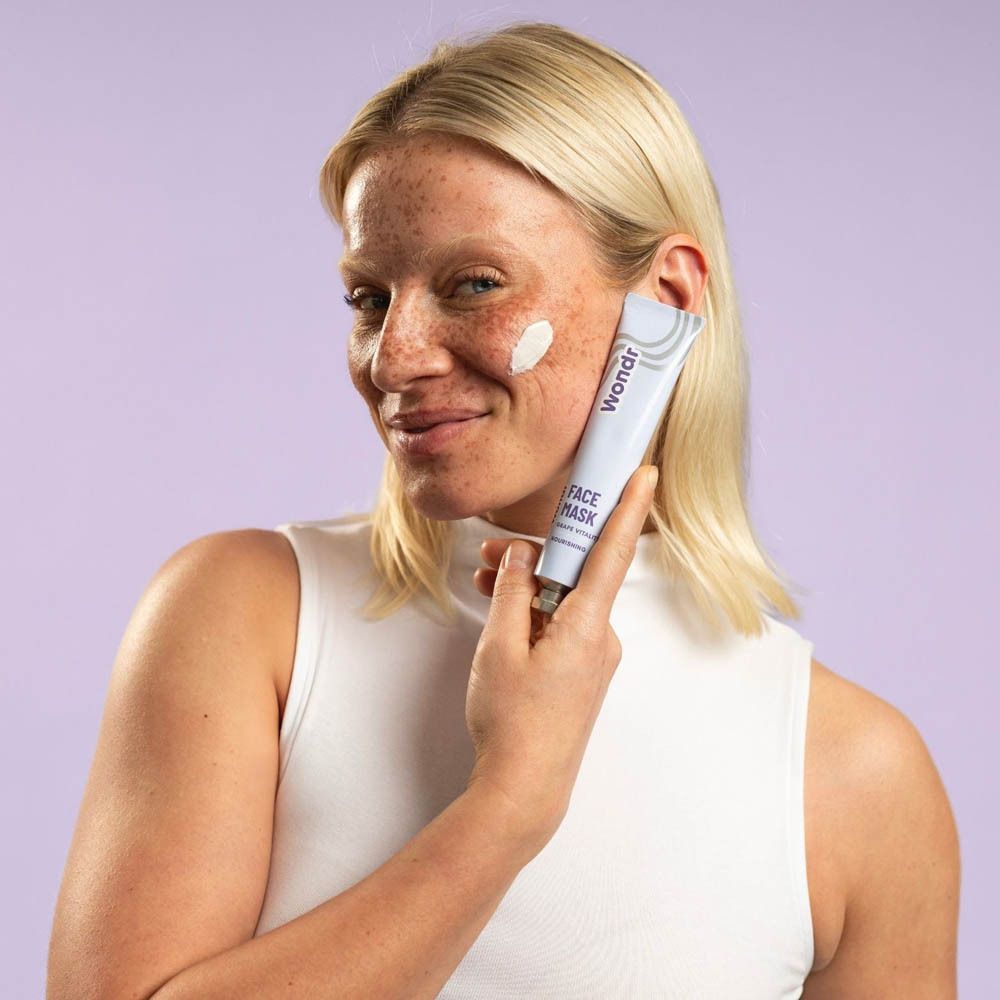 Vrouw houdt tube vast. Tube met opschrift: Wondr, Face Mask, Grape Vitality, Nourishing. Crème op gezicht.