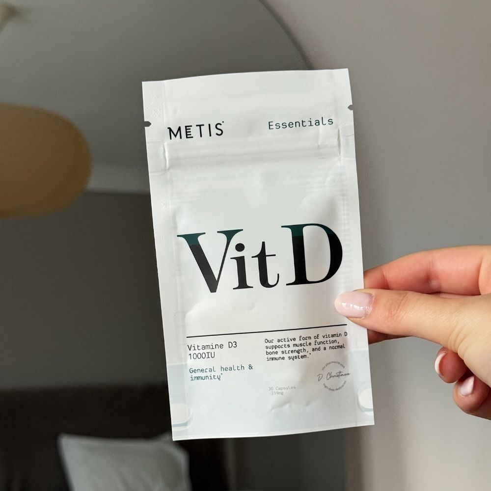 Hand houdt een verpakking van Metis Essentials Vit D vast. Opschrift: Vit D, vitamine D3 1000IE.