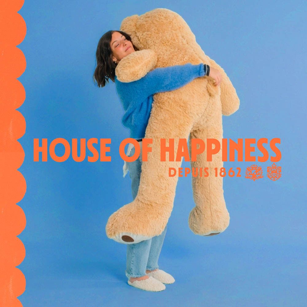 Femme étreignant un ours en peluche. Texte: House of Happiness. Flacon: Mandarine Doudou. Orange.