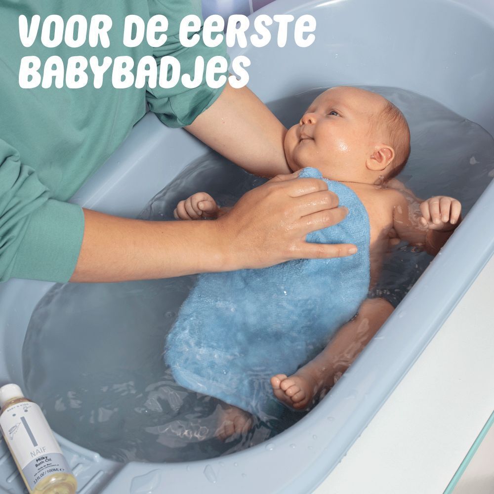 Baby in bad. Persoon wast baby met blauwe doek. Naïf Badolie fles aan de rand.