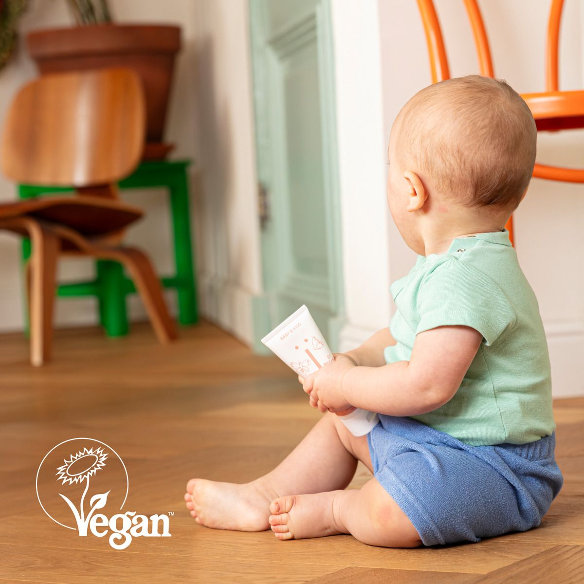 Baby zit op de grond en houdt een tube babycrème vast. Tekst: Vegan.