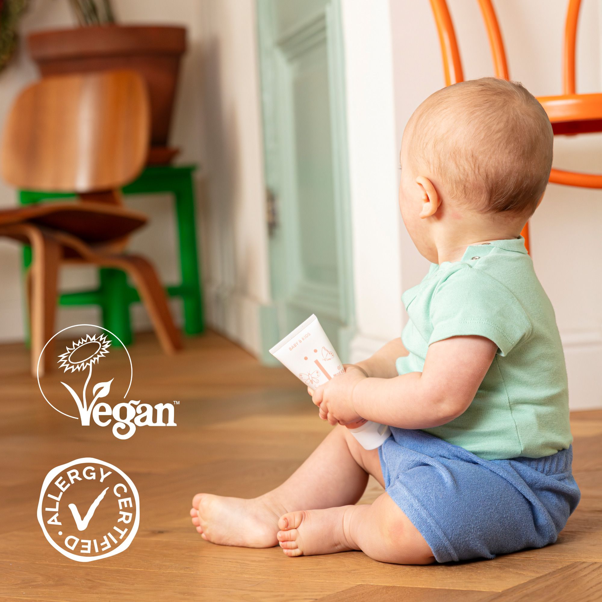 Bébé assis par terre tenant un tube. Logo végan et certification allergie. Plancher en bois.