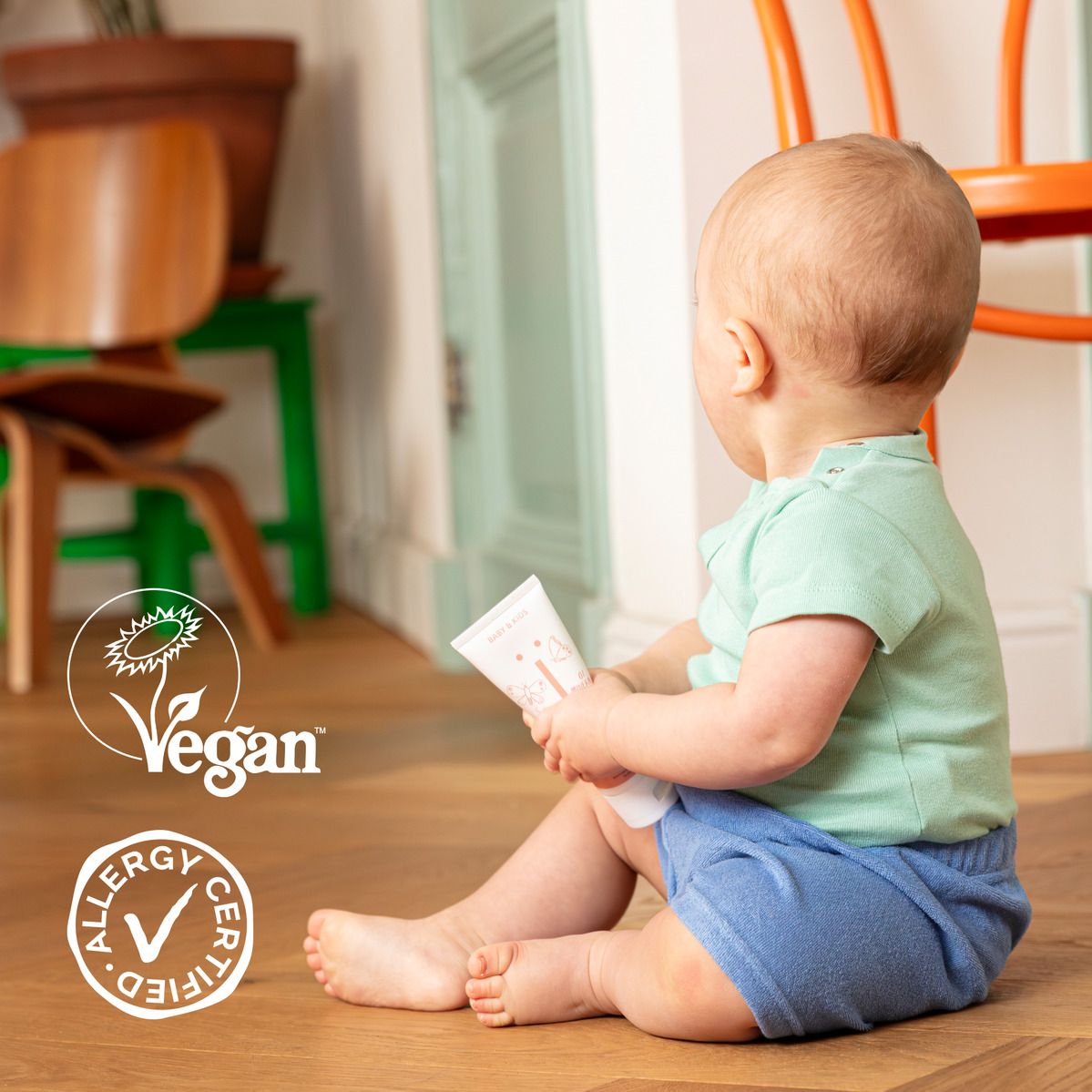 Baby zit op de grond met een tube. Vegan logo en allergie certificering. Houten vloer.