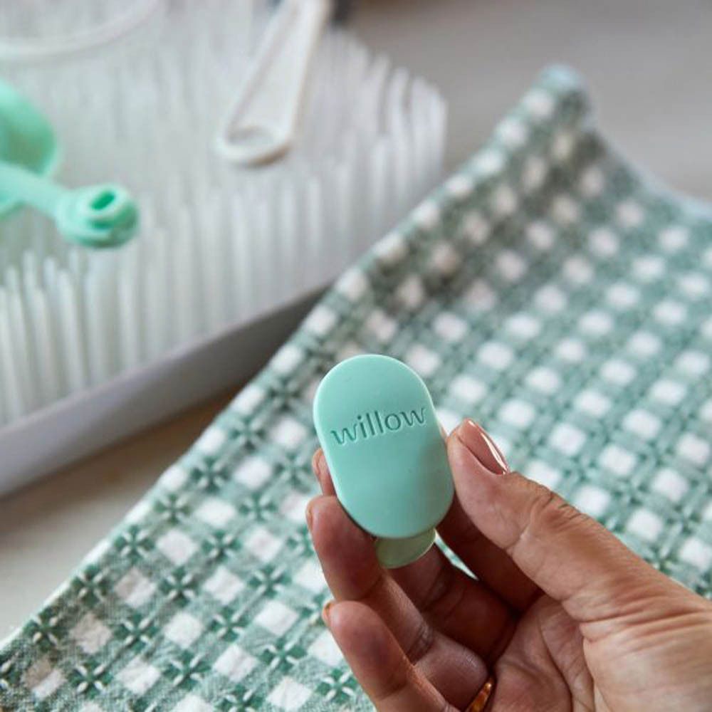 Een hand houdt een mintgroen Elvie Wave ventiel met 'willow' logo vast. Op de achtergrond een afdruiprek en een handdoek.