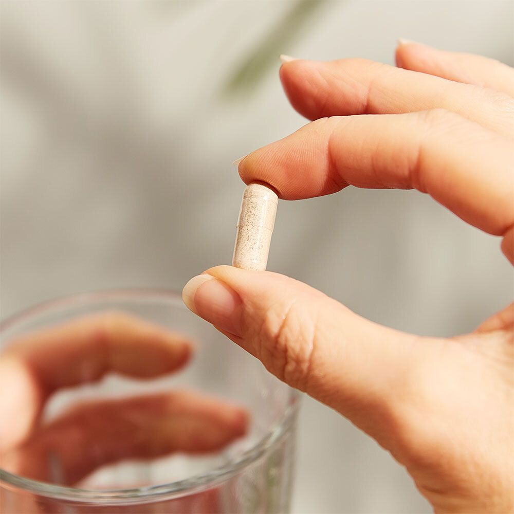 Une main tient une gélule au-dessus d'un verre d'eau. La gélule est beige.