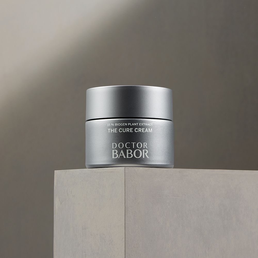 Zilverkleurige crèmepot op een grijs oppervlak. Opschrift: THE CURE CREAM, DOCTOR BABOR.