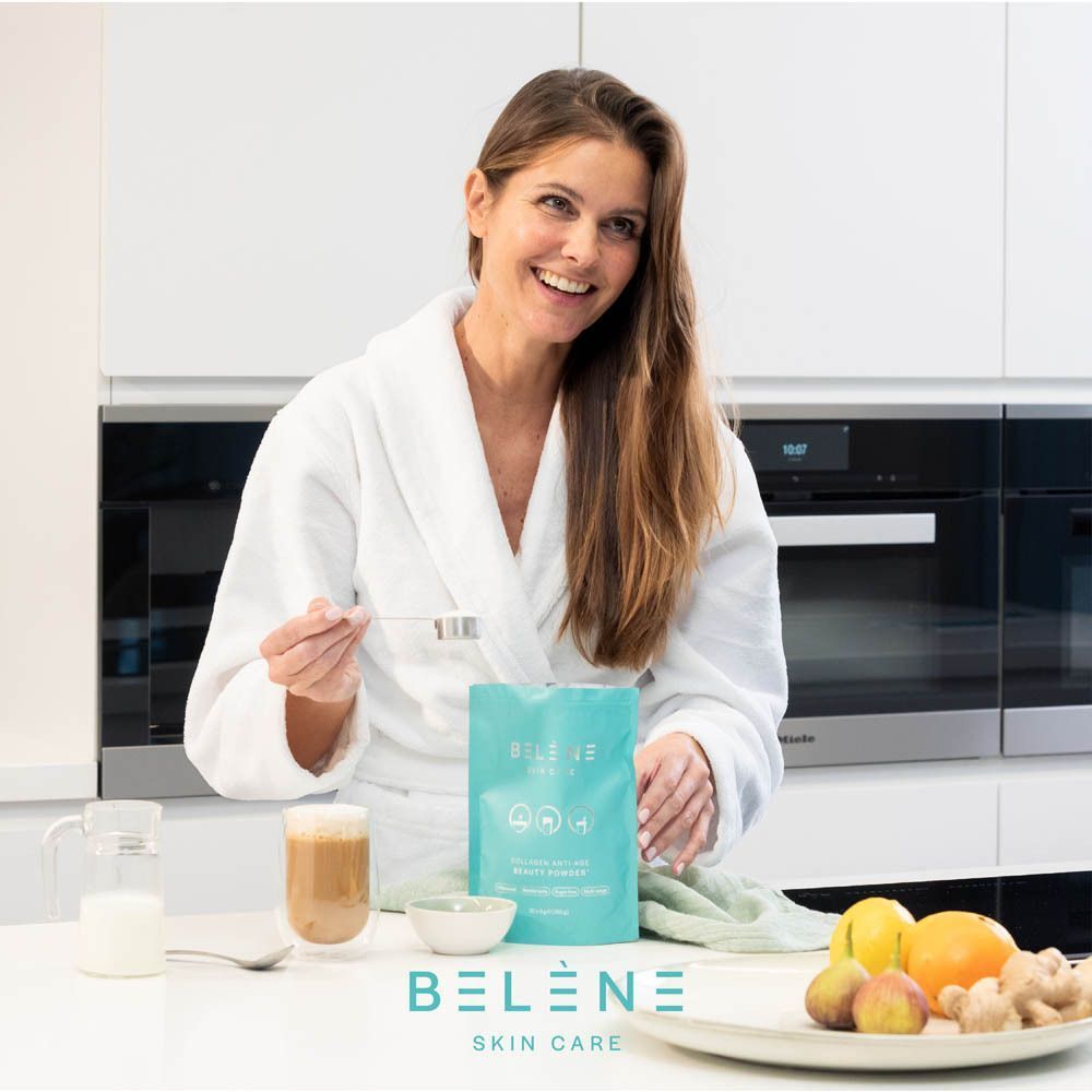 Femme en peignoir dans une cuisine. Tient une cuillère et un sachet Belène Skin Care. Sur la table : boisson, fruits, gingembre. Logo visible.