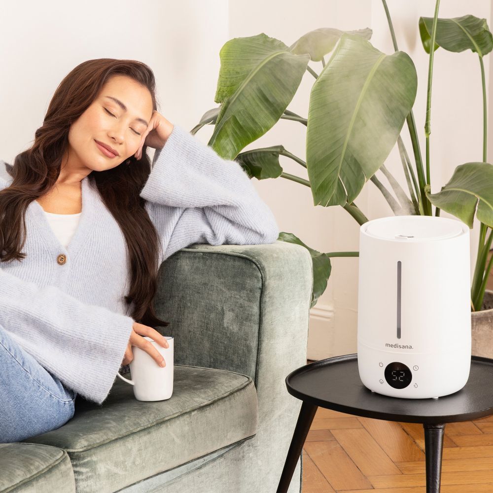 Une femme est assise à côté d'un humidificateur blanc. L'appareil a un affichage numérique et le logo "medisana".