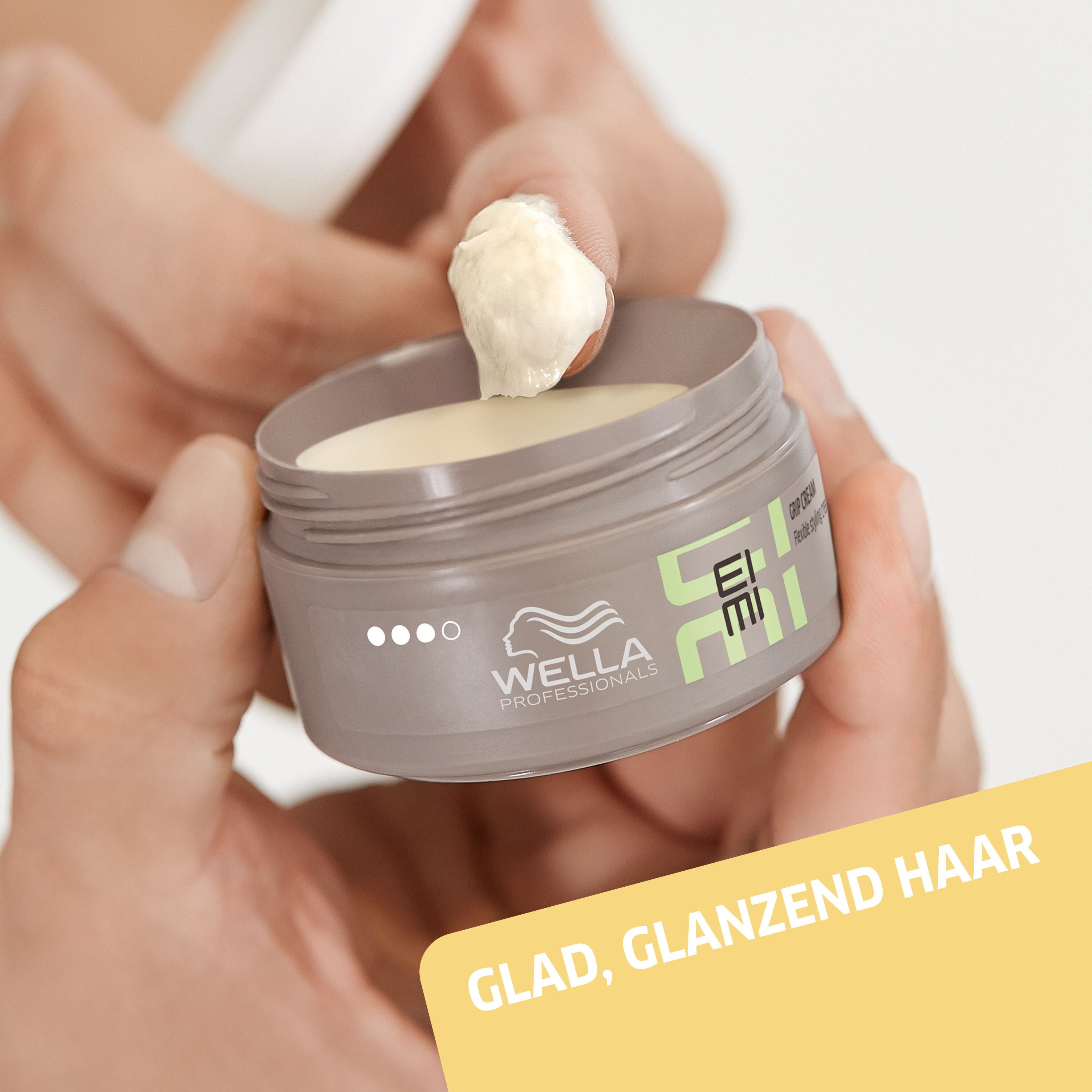 Handen houden een pot pommade vast. Opschrift: EIMI. Tekst: GLAD, GLANZEND HAAR.