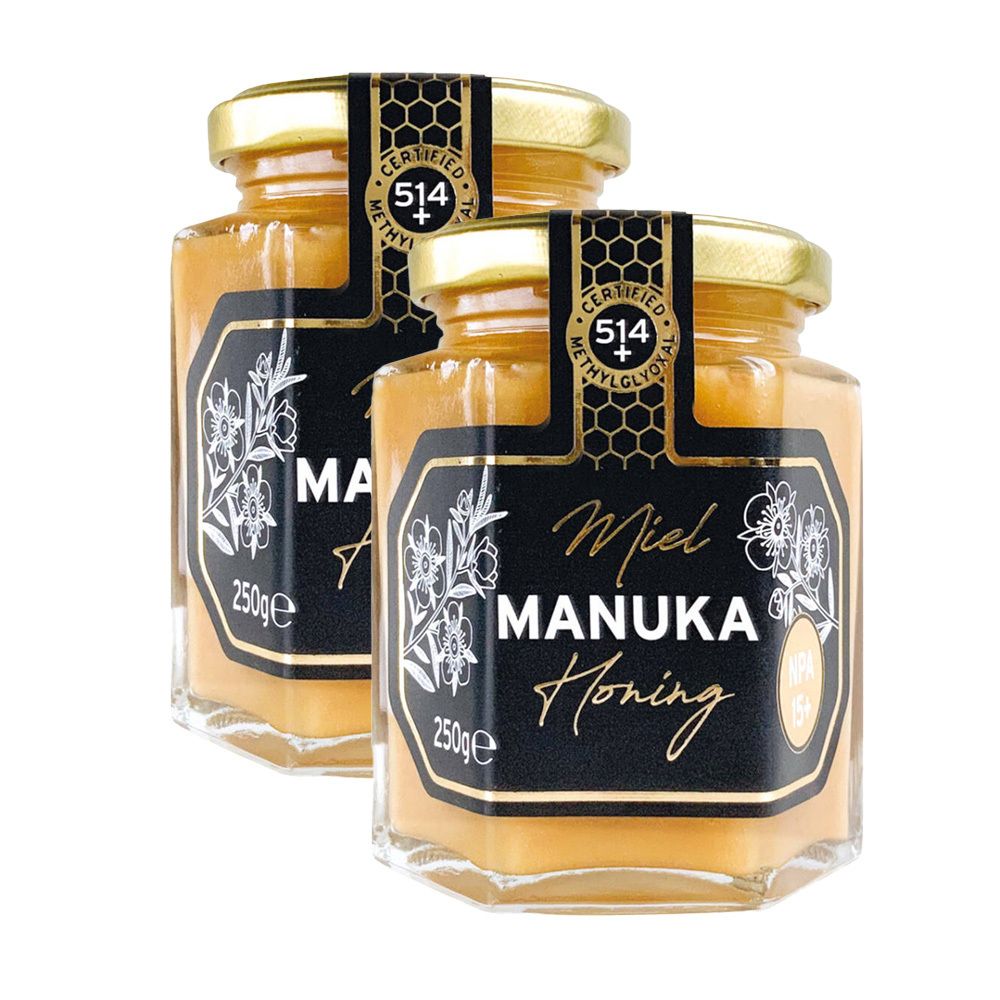 Twee potten honing. Zwarte etiketten met witte tekst: Miel Manuka Honing. Gouden deksels. Certificeringsstickers.