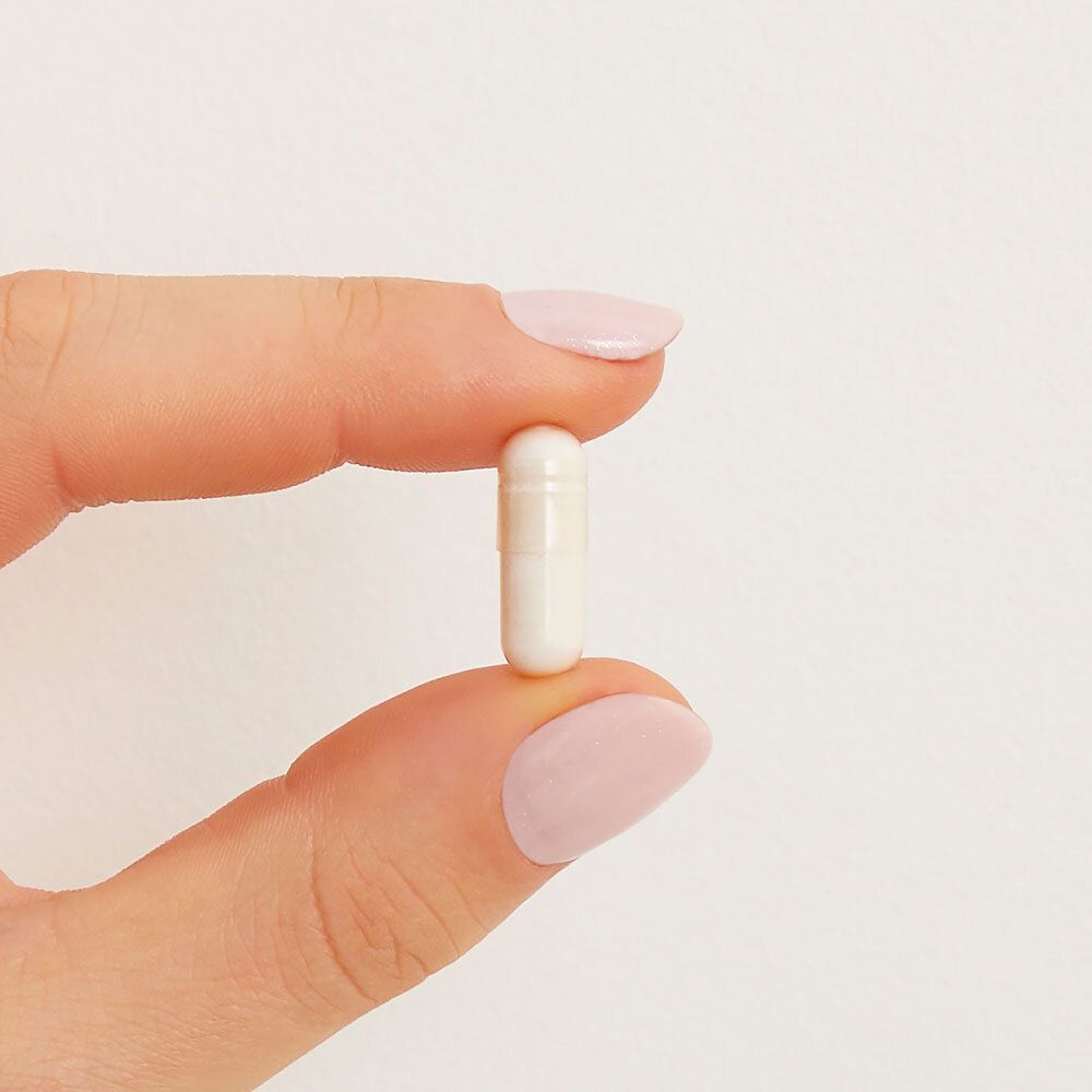 Een witte capsule, vastgehouden door een hand met roze gelakte nagels. De capsule is langwerpig.