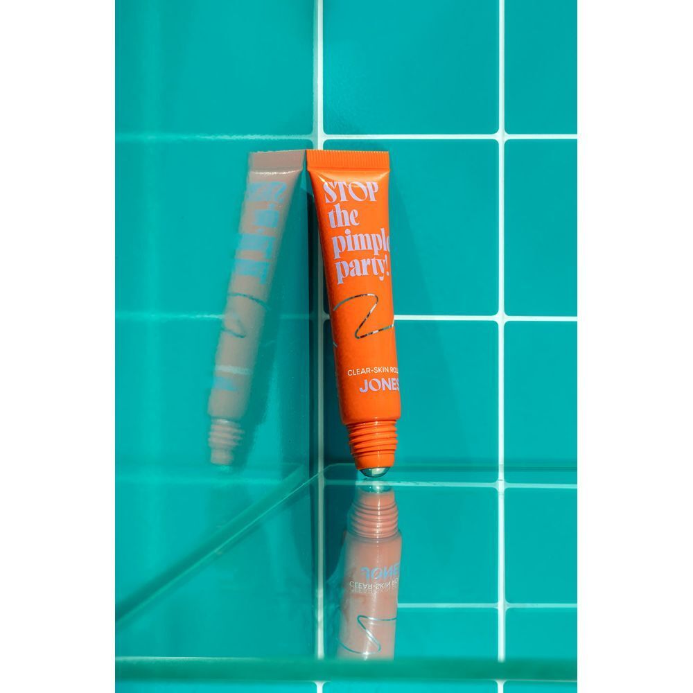 Oranje tube met tekst. Tekst: Stop the Pimple Party! Clear-Skin Roller. Merk: JONES. Reflectie.