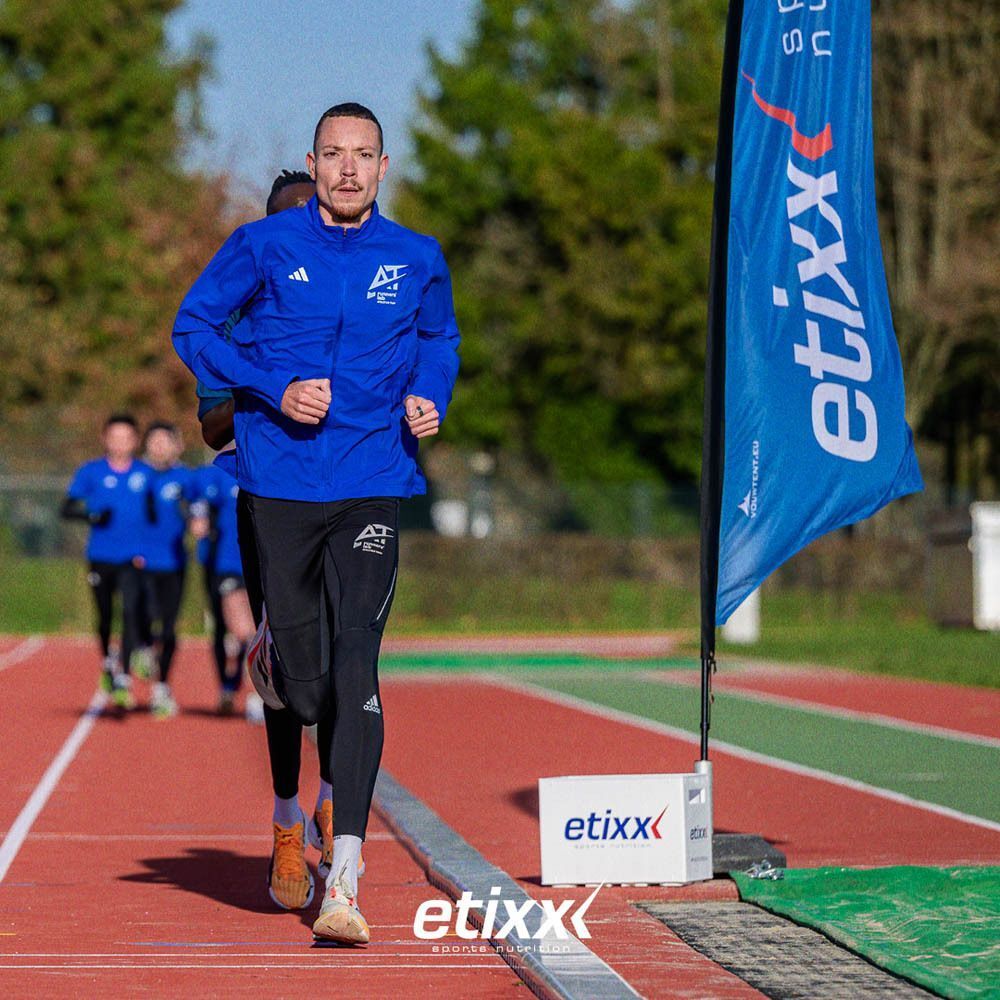 Coureur en veste bleue. Autres coureurs en arrière-plan. Bannière Etixx et logo Etixx au sol.