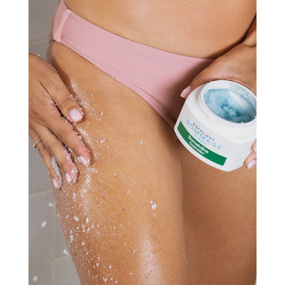 Persoon wrijft peeling op been. Pot met peeling zichtbaar. Opschrift: EXFOLIANT GOMMAGE, Somatoline Cosmetic.