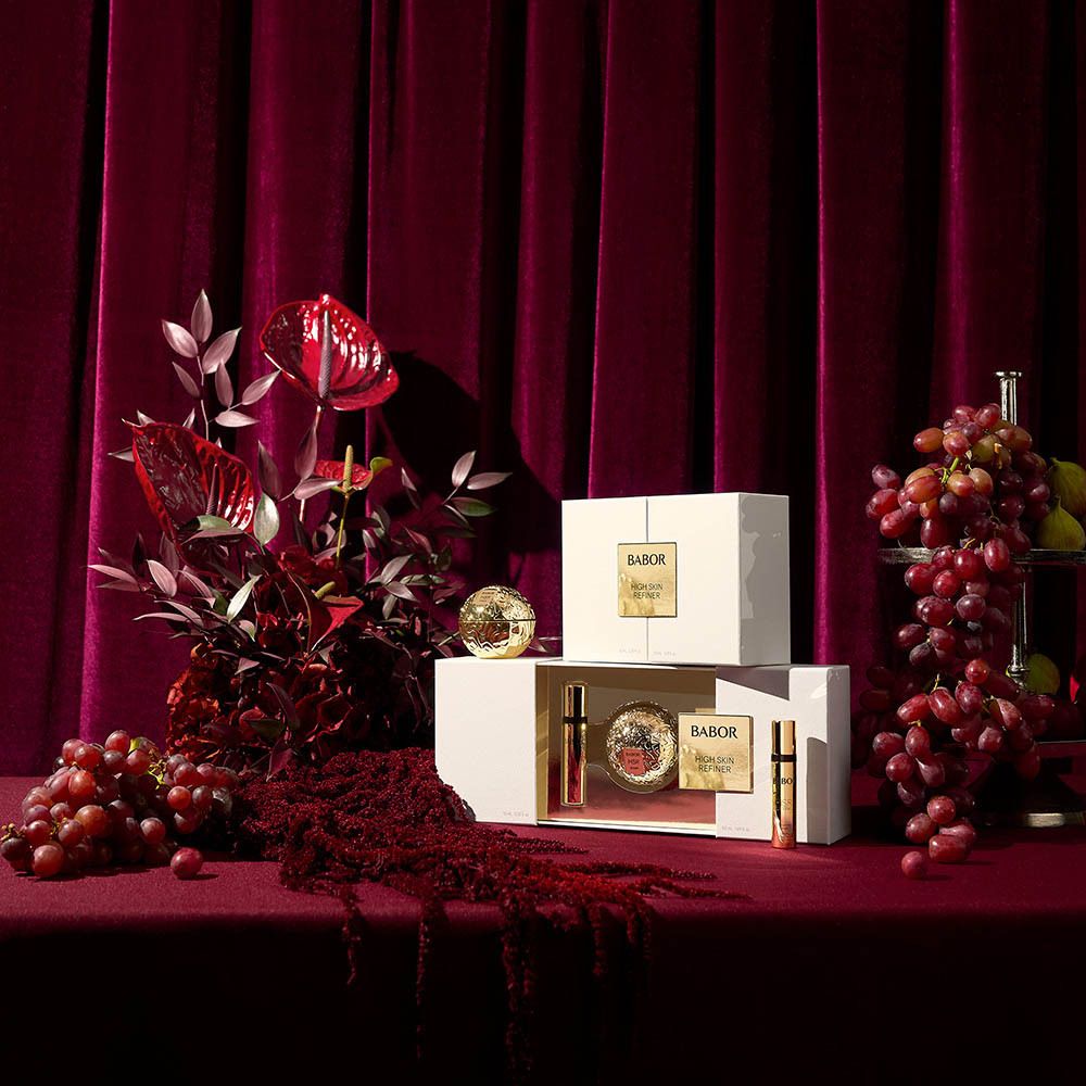 Productpresentatie met geschenkverpakkingen en decoratie. Opschrift: BABOR HIGH SKIN REFINER. Producten op rode tafel.