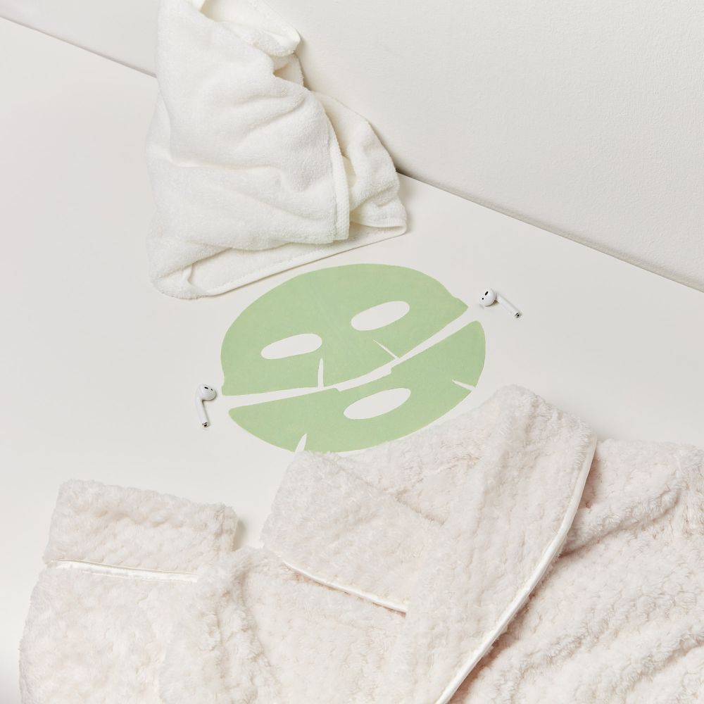 Masque facial, serviette et peignoir sur une surface blanche. Le masque est vert clair.