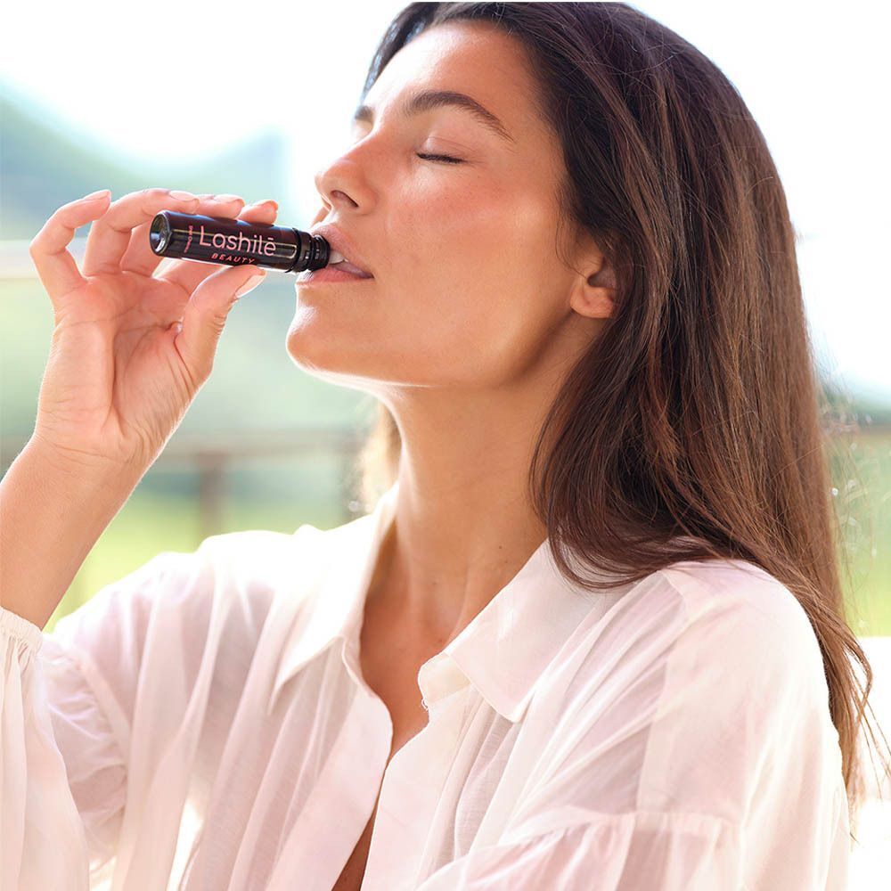 Vrouw drinkt een zwarte shot met Lashilé Beauty. Tekst: Good Hair Shots. Achtergrond: fruit.