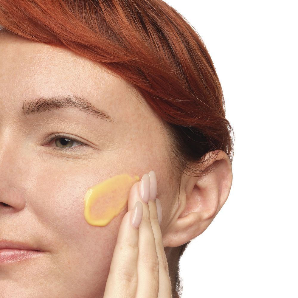 Een hand brengt gele emulsie op de huid aan. Een deel van het gezicht is zichtbaar.