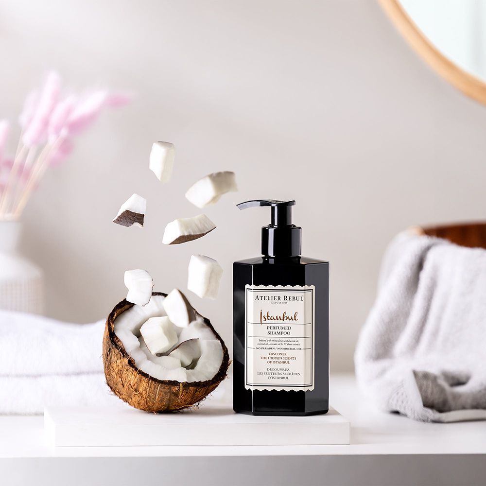 Flacon noir avec pompe doseuse, à côté d'une noix de coco et de morceaux de noix de coco. "ATELIER REBUL Istanbul Shampooing Parfumé".