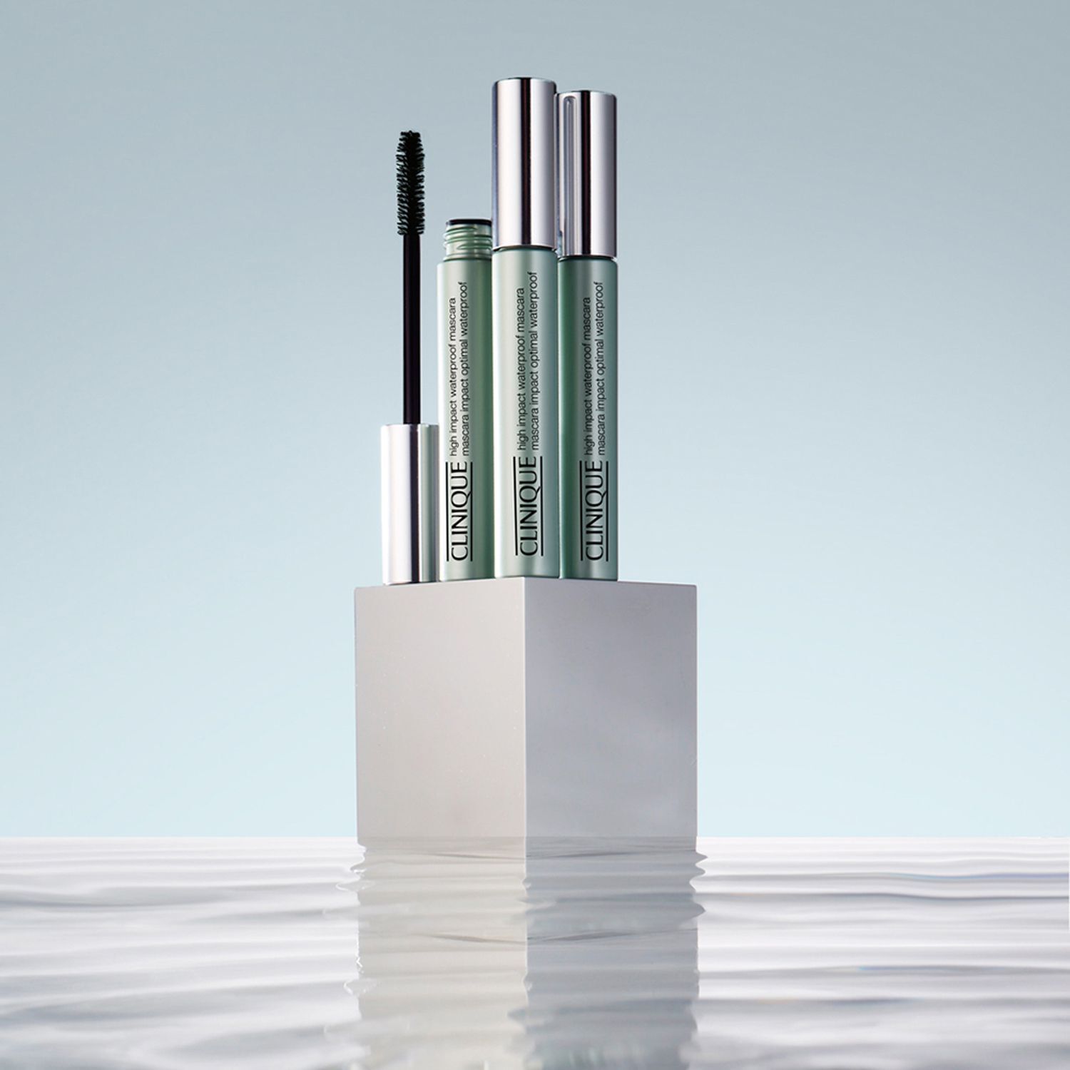 Drie CLINIQUE High Impact Waterproof mascara tubes op een wit voetstuk. Achtergrond: water met reflecties.