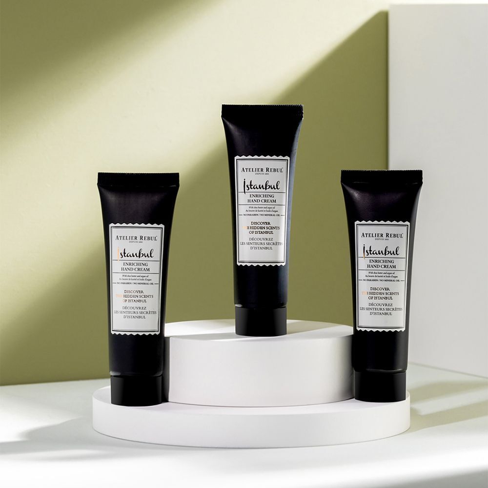 Drie zwarte tubes handcrème op een wit voetstuk. Opschrift: ATELIER REBUL Istanbul Enriching Hand Cream.