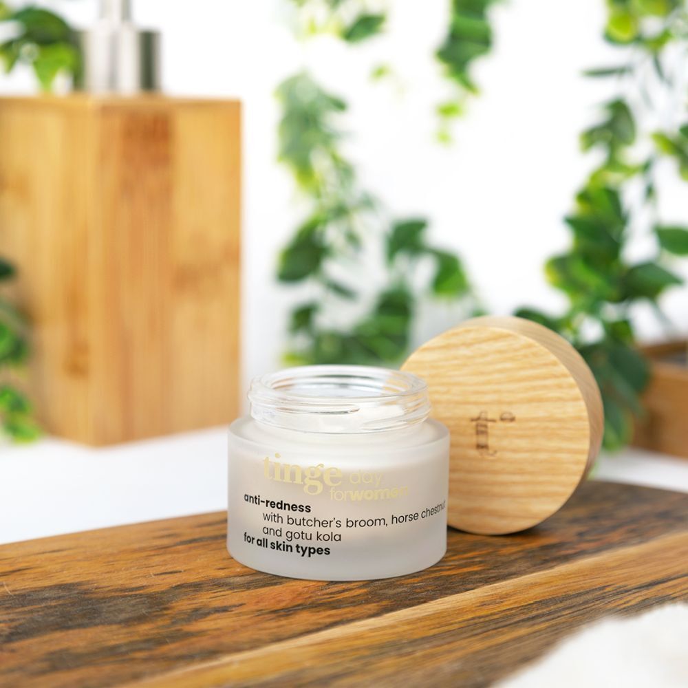 Potje crème anti-roodheid met houten deksel, op houten plank. Houten zeepdispenser en groene planten ernaast. Tekst: anti-redness, for all skin types.