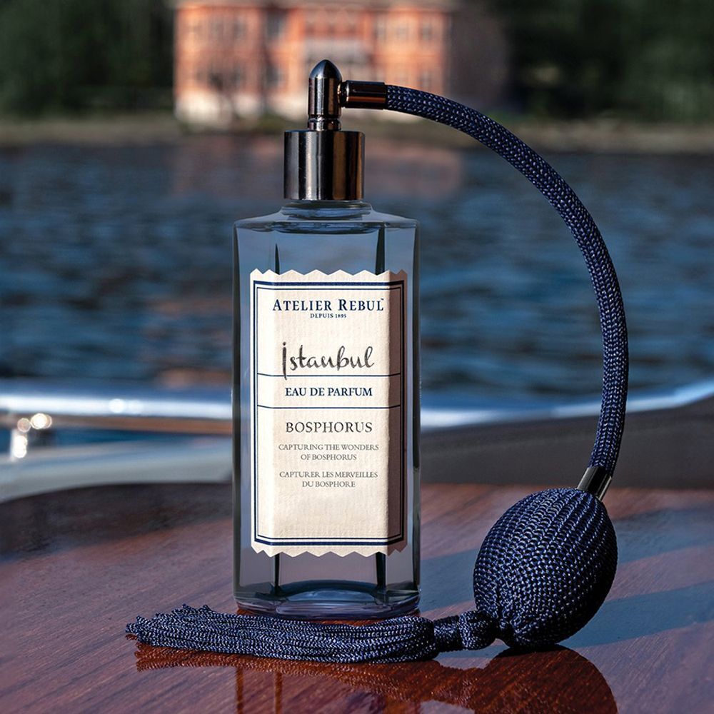 Donkerblauwe fles met verstuiver. Opschrift: Istanbul Bosphorus Eau De Parfum. Fles op hout. Achtergrond: water.
