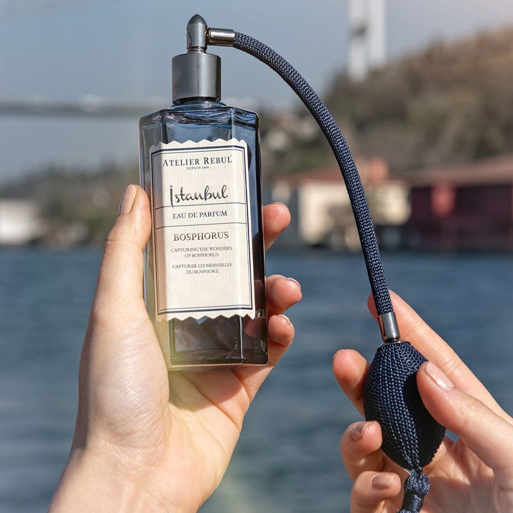 Handen houden een donkerblauwe fles met verstuiver. Opschrift: Istanbul Bosphorus Eau De Parfum. Achtergrond: water en brug.