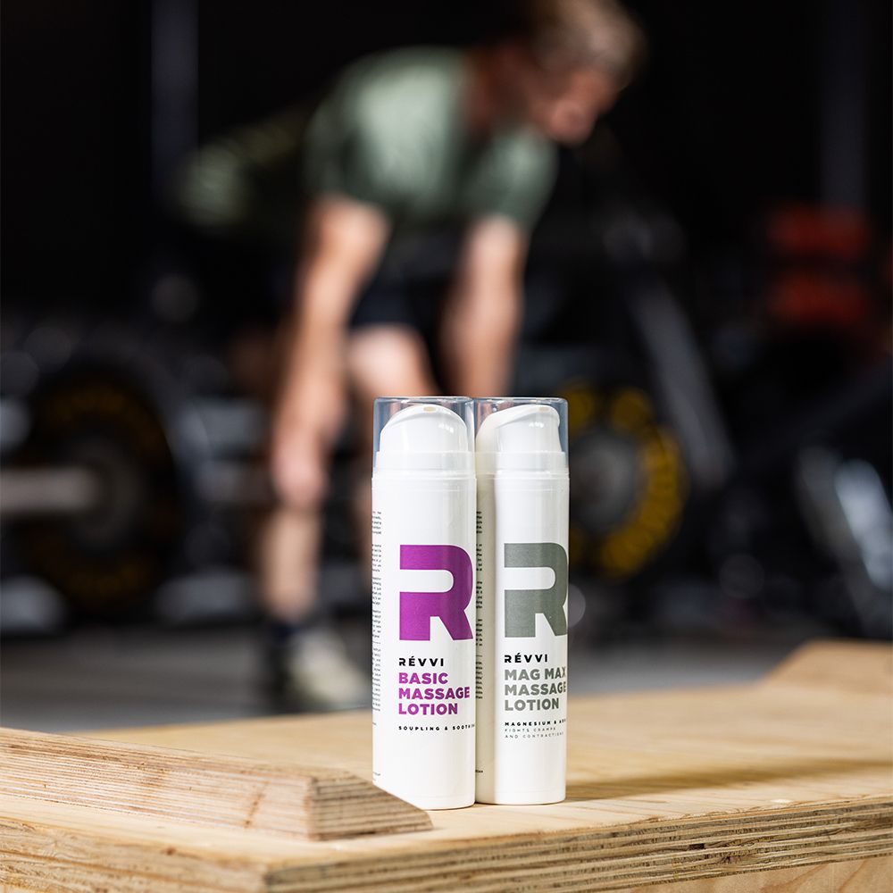 Twee witte flessen met roze logo en tekst: RÉVVI Basic Massage Lotion. Op houten oppervlak.