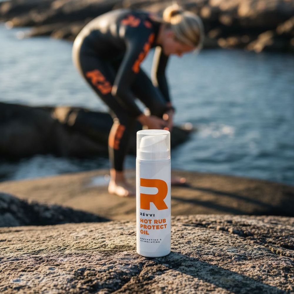 Flacon blanc avec logo "R" orange. Texte : RÉVVI, HOT RUB PROTECT OIL. Bouchon transparent. Personne à la mer.