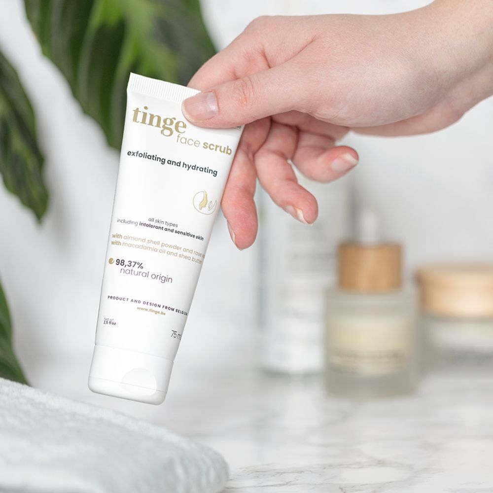 Main tenant un tube de gommage visage Tinge. Blanc, avec texte et logo. Arrière-plan flou.