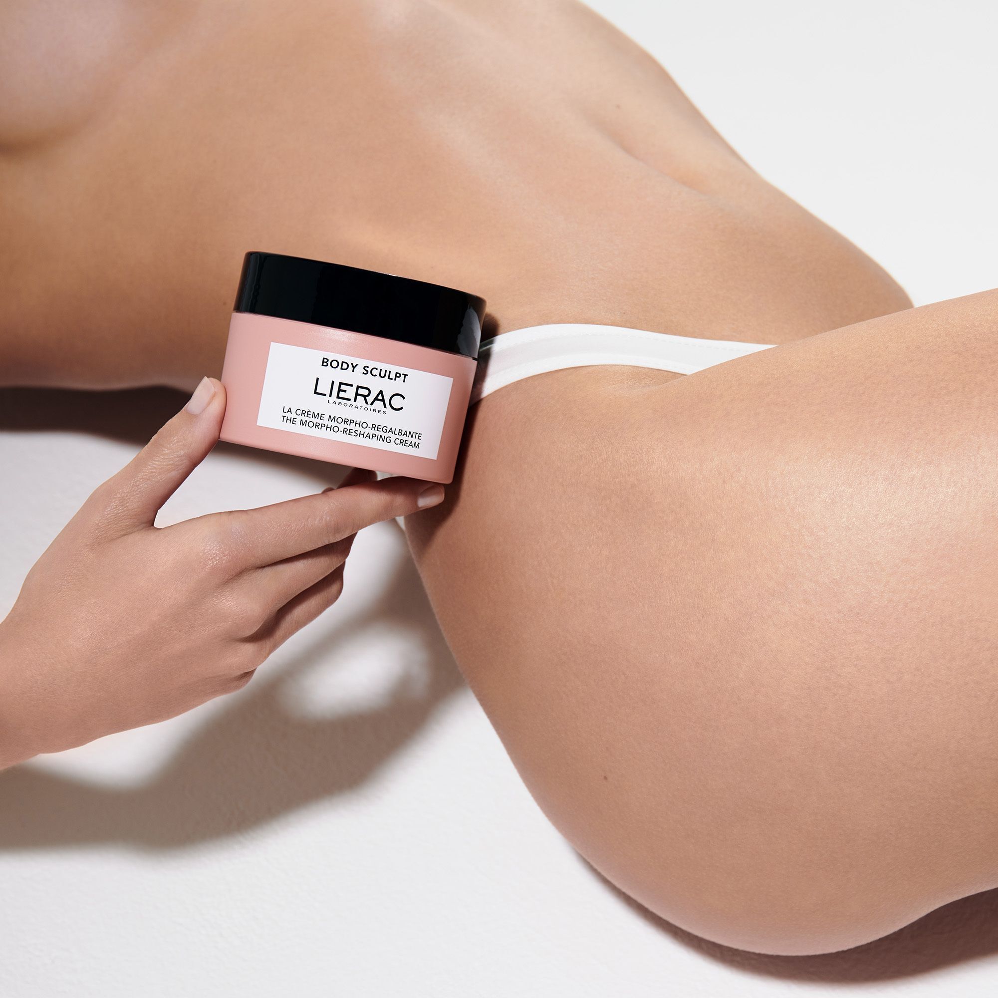 Hand die een roze pot met zwart deksel vasthoudt. Opschrift: BODY SCULPT, LIERAC, LA CRÈME MORPHO-REGALBANTE, THE MORPHO-RESHAPING CREAM.