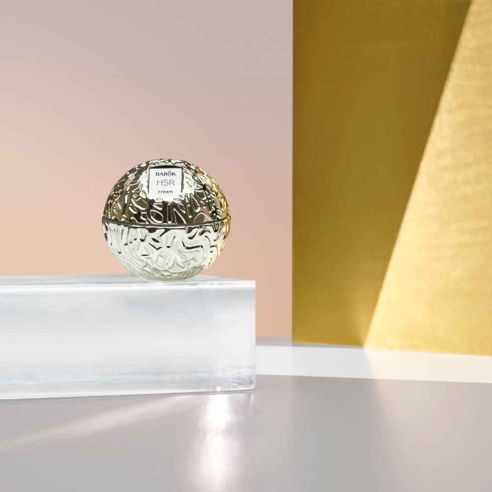 Goudkleurige crèmetiegel met Babor HSR cream op een glazen plaat. Achtergrond: goud en roze.