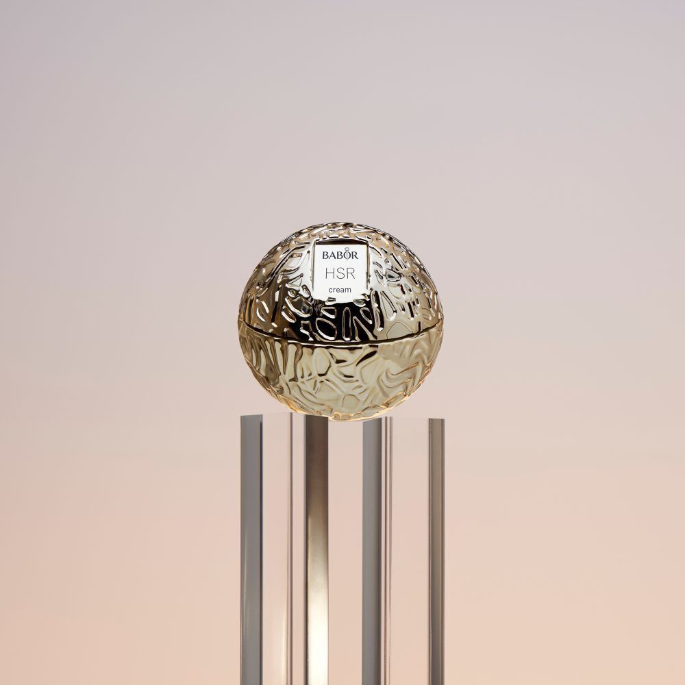 Goudkleurige crèmetiegel met Babor HSR cream op een glazen plaat. Achtergrond: roze en goud.