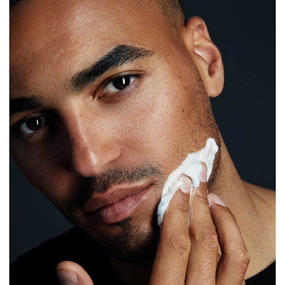 Un homme applique de la crème à raser. La crème est blanche. L'homme a une barbe et la peau foncée.