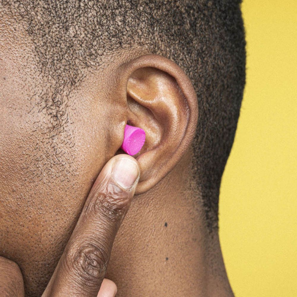 Personne insérant un bouchon d'oreille rose dans une oreille. Fond jaune. Un seul bouchon d'oreille visible.