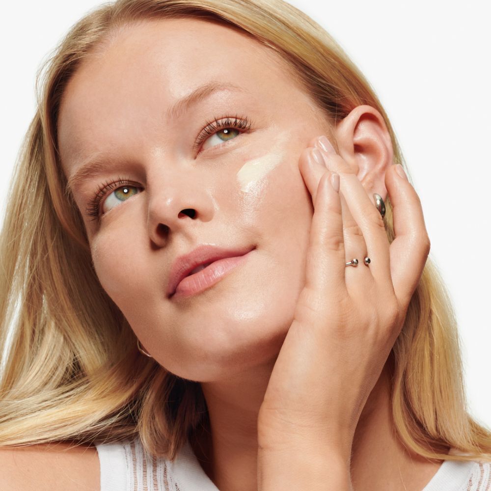 Femme appliquant de la crème sur son visage. Elle touche sa joue avec sa main. Peau claire, cheveux blonds.