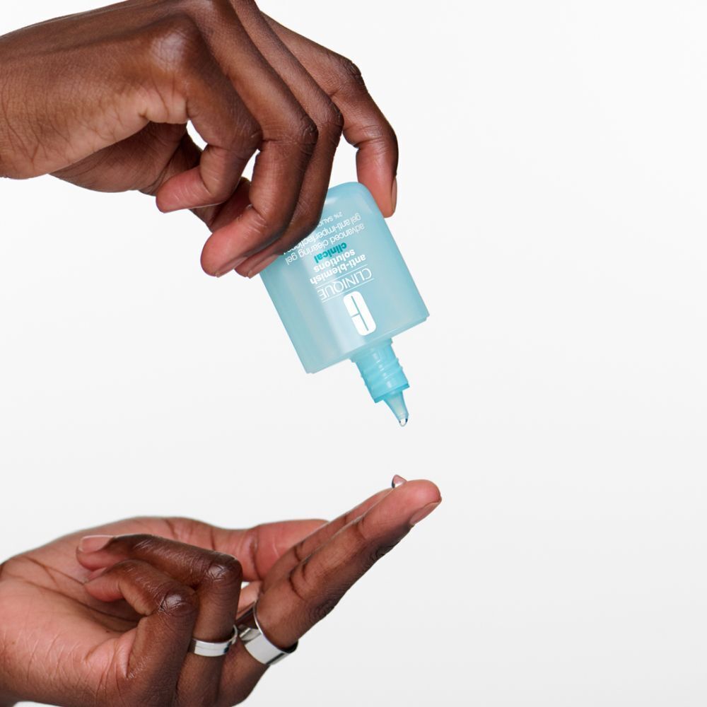 Mains tenant un flacon de gel. Le gel coule sur un doigt. Flacon : bleu clair, bouchon blanc, logo Clinique.