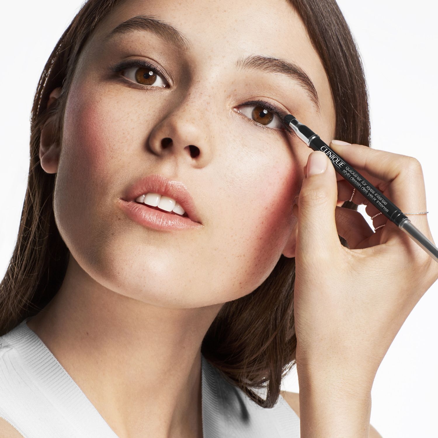Une jeune femme avec des taches de rousseur applique de l'eye-liner noir avec un crayon Clinique.
