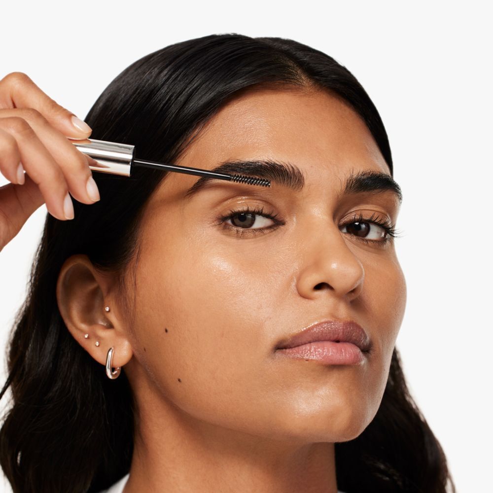 Une femme brosse ses sourcils avec un gel pour sourcils. Le produit n'est pas entièrement visible. Marque: CLINIQUE.