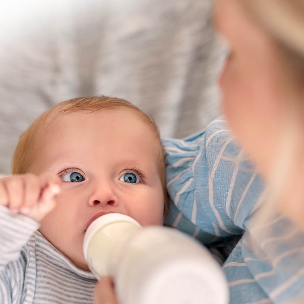 Un bébé boit dans un biberon blanc, tenu par une personne.