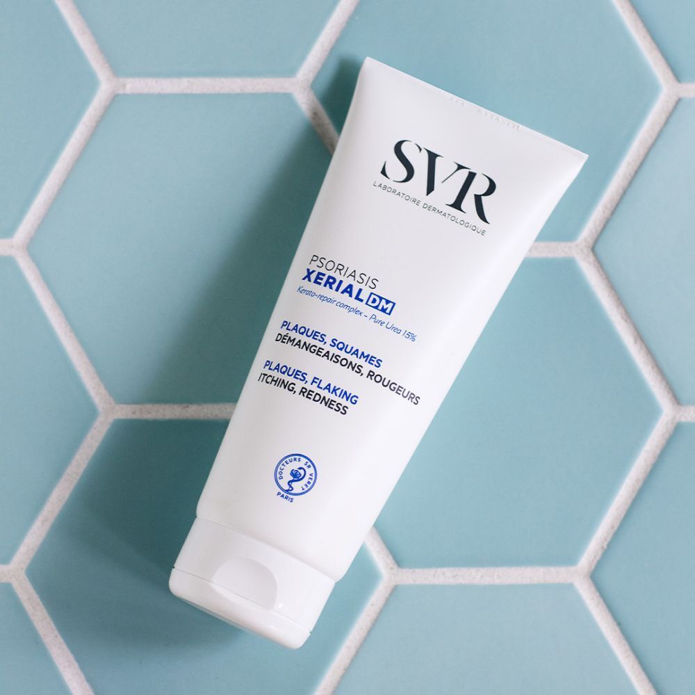 Tube blanc SVR Xerial DM Psoriasis sur fond bleu hexagonal. Texte: Plaques, Squames, Démangeaisons, Rougeurs. Logo SVR.